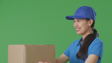 close up of asian female courier in blue uniform giving a carton to someone and smiling while delivering it on green screen background in the studio