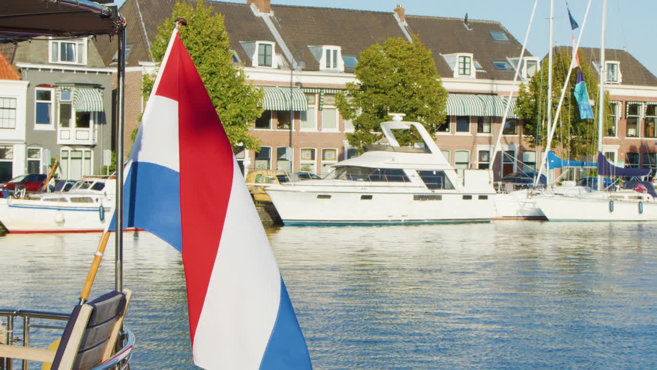 Dutch flag waving beside canal with historic buildings, boats, and blue sky in Haarlem