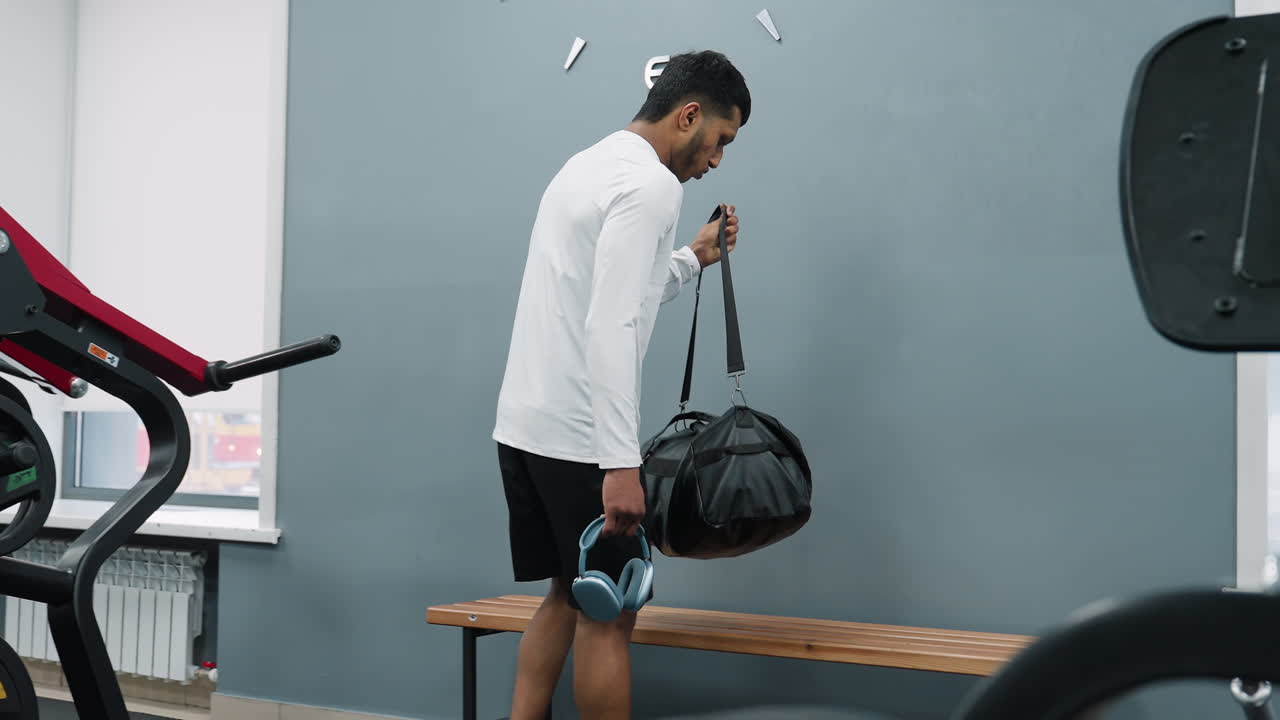young man in white jacket and black shorts sets leather gym bag on wooden bench beside red exercise machine inside bright fitness center corridor while sunlight reveals car passing by through window
