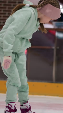 una vista cercana de un niño en un traje verde menta y una cinta rosa inclinándose para limpiarse en una pista de hielo, el fondo muestra una vista borrosa de otras personas y equipo de patinaje sobre hielo