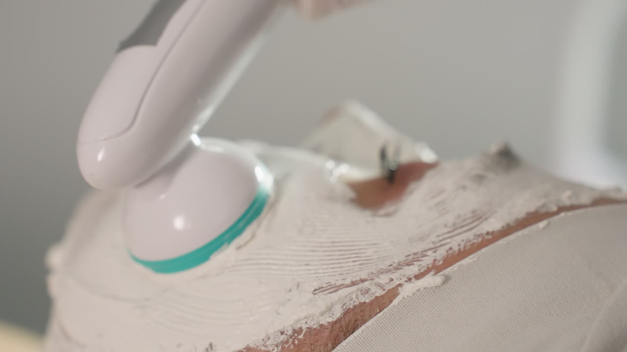 Skin care specialist uses electric facial massager vibrating in circular motions to smooth thick cream mask on client face under bright spa light for deep skin rejuvenation and relaxation