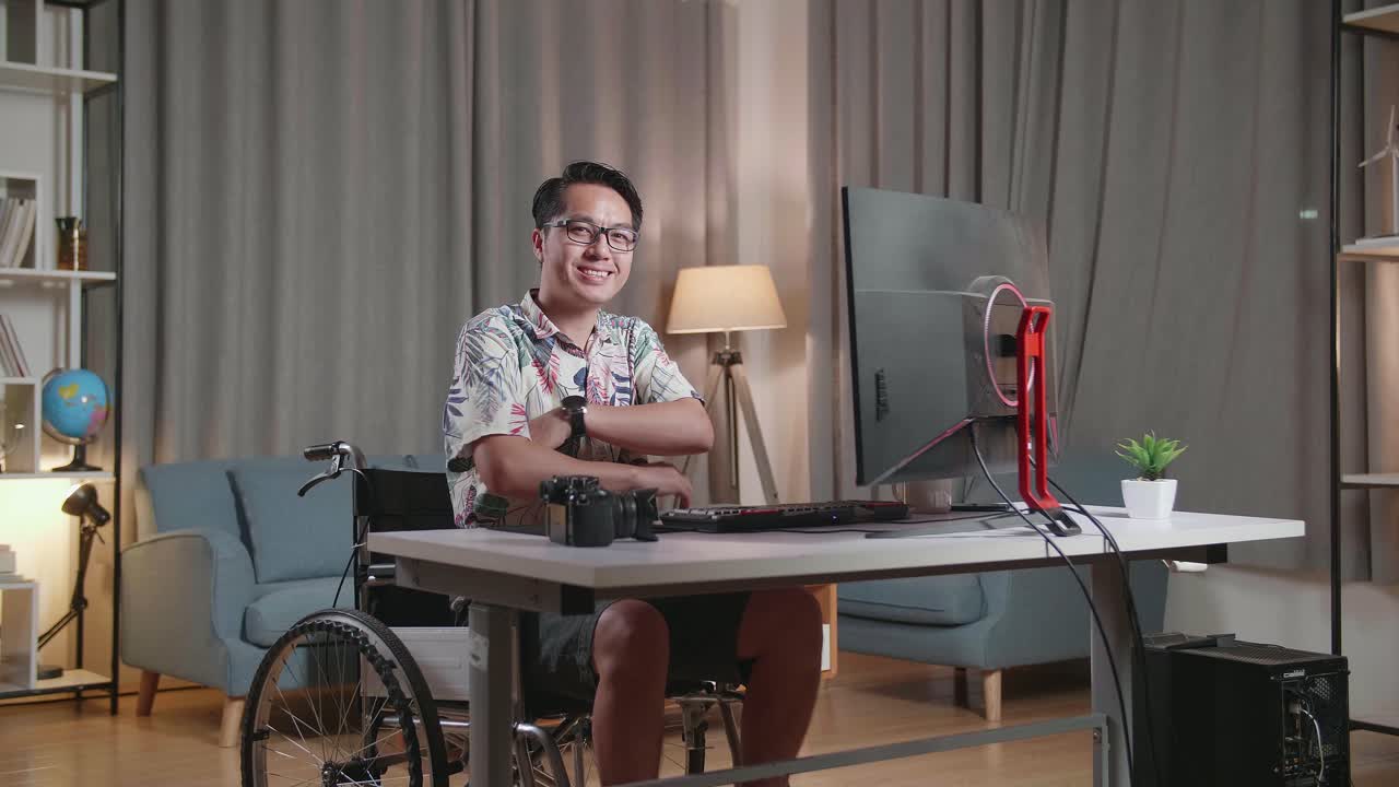hombre asiático en silla de ruedas sonriendo y cruzando los brazos a la cámara mientras edita el video por un escritorio al lado de la cámara en casa