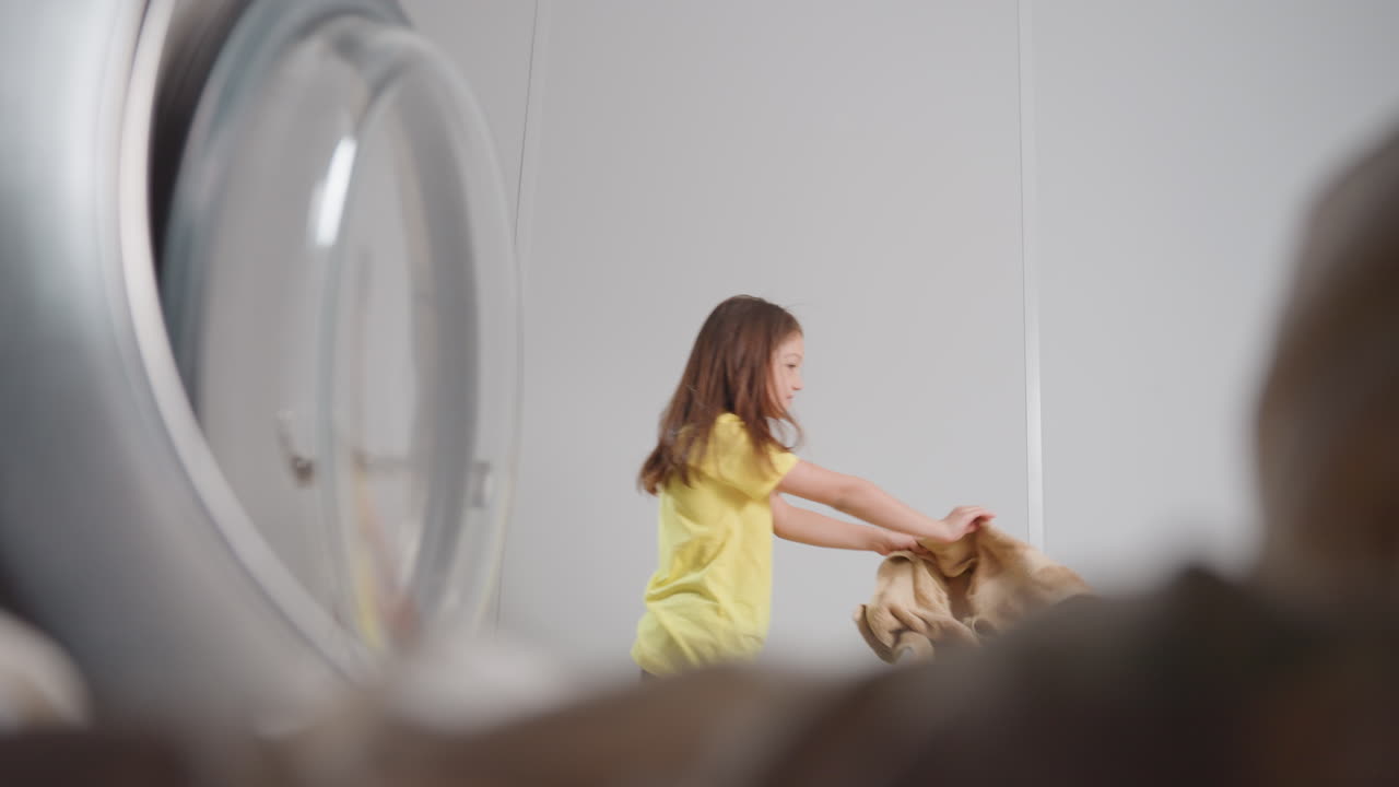 Inside washer view shows active kid with towel dancing backward past open door, round glass rim framing scene, soft motion blur, bright laundry room, playful energy, clean fabrics, captured from drum