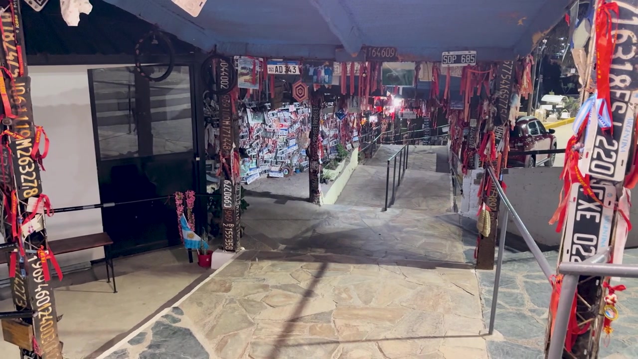 Panoramica statica a destra che inquadra l'esterno illuminato del Santuario della Difunta Correa a Vallecito di notte, mostrando fasce di preghiera appese, offerte votive e decorazioni tradizionali da pellegrinaggio.