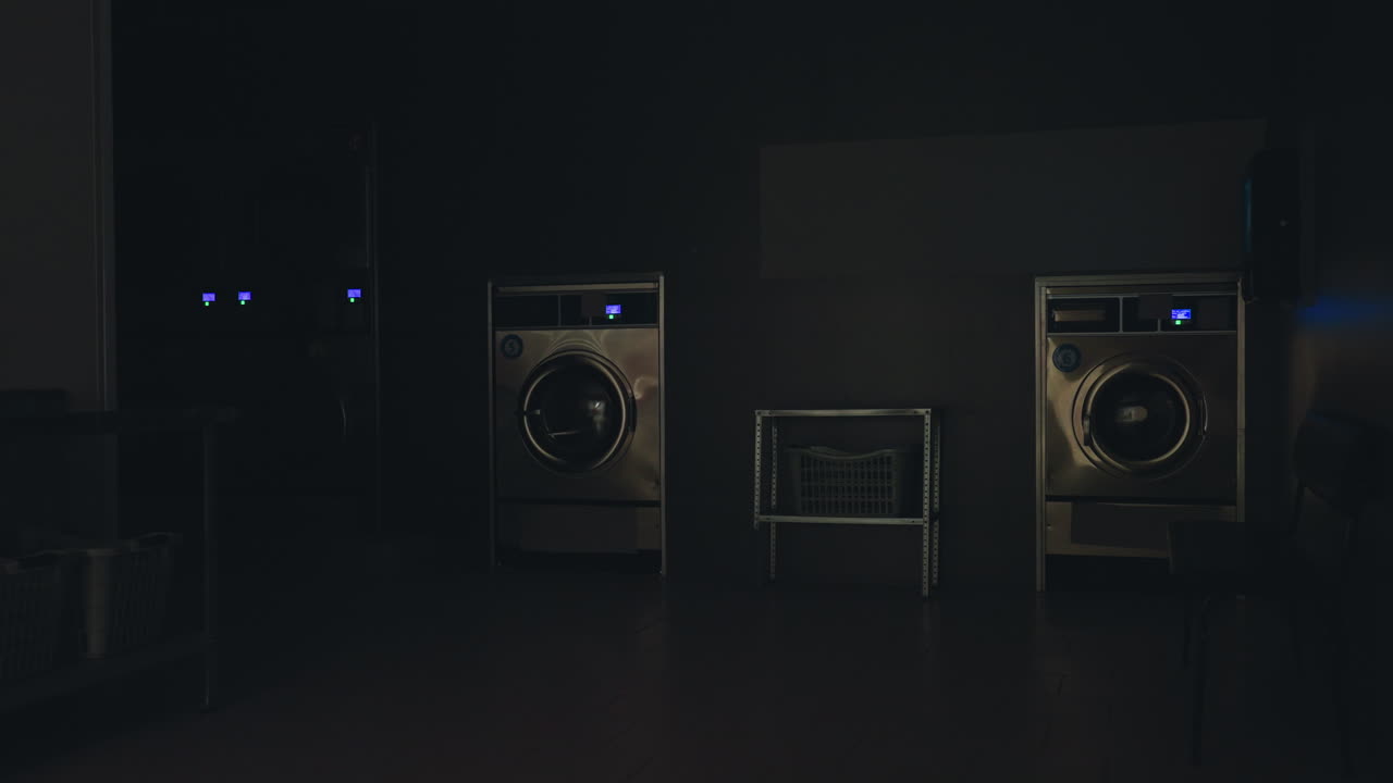 Bright modern laundry shop interior with stainless washing machines, organized laundry baskets, black bench, and tiled floor as overhead lights switch off, creating calm service facility environment
