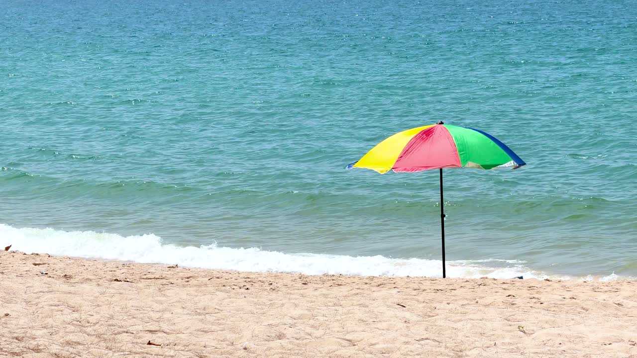 Plan large fixe d'un parasol arc-en-ciel éclatant sur du sable doré avec des vagues d'océan turquoise
