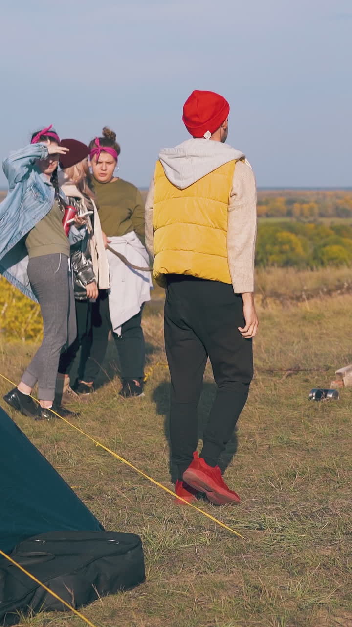happy girls look at handsome guy going out of tent in comfortable camp on river bank against blue sky in morning