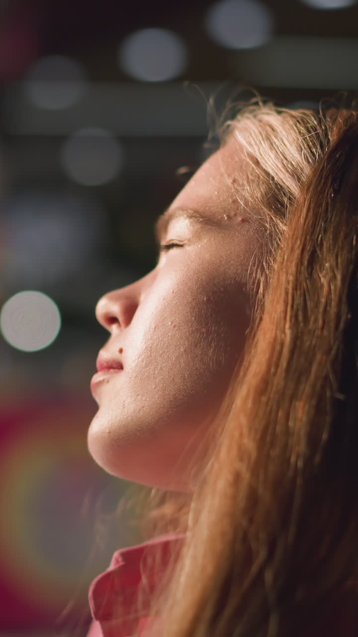 una dama blanca con un vestido rosa se relaja en una silla de madera, la cara muestra espinillas ligeras, mientras que las luces borrosas crean un efecto brillante en el fondo, las personas son visibles pasando en el fondo borroso
