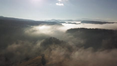 High-flight-above-mountains-and-valleys-covered-with-white-carpet-clouds-with-bright-white-sun-rays-in-background,-aerial-overhead-pull-back