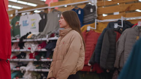 a girl with long brown hair, wearing a light brown jacket, is seen standing and looking around thoughtfully in a clothing store. various jackets and shirts are displayed on racks in the background