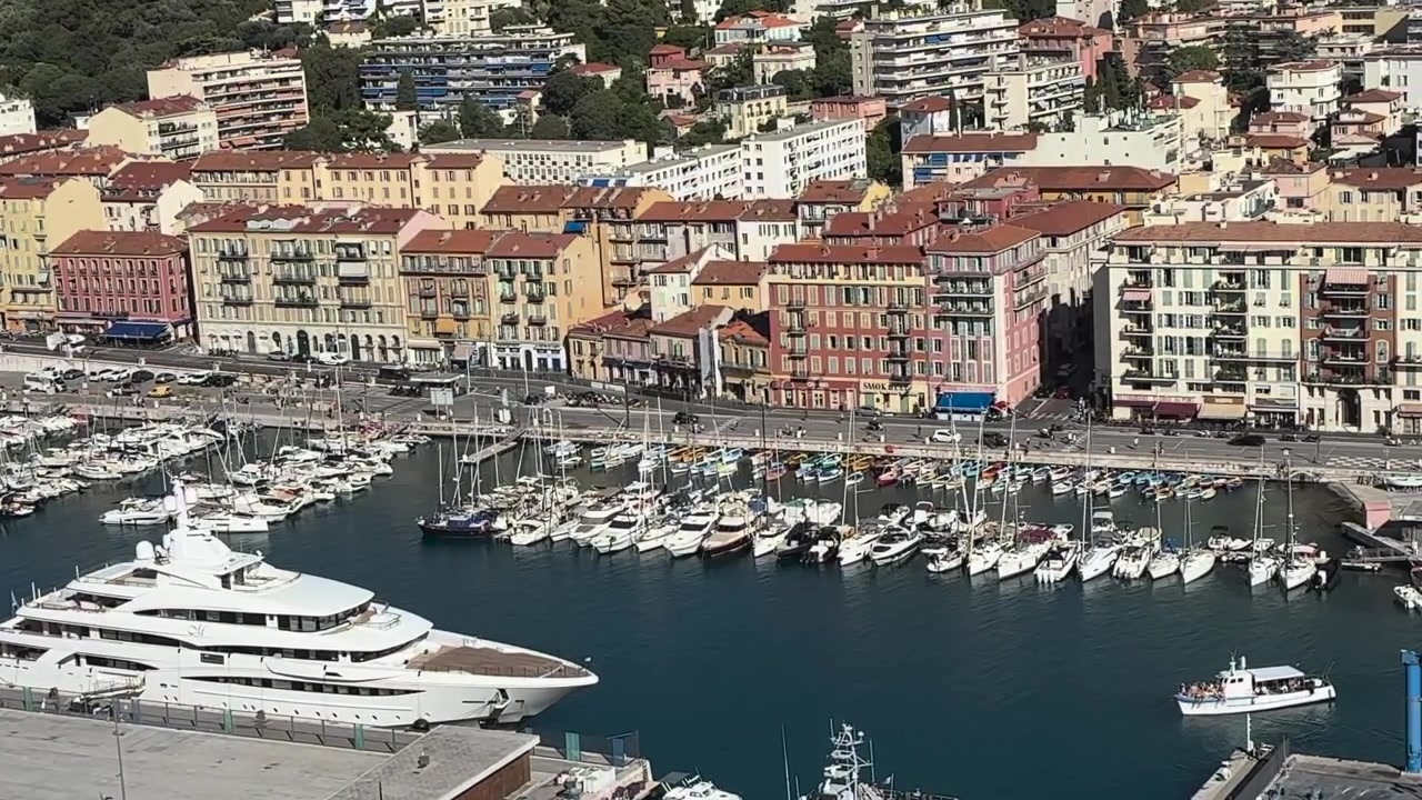 Vista dall'alto di un superyacht di lusso e file di barche a vela nel Porto di Nizza, con sullo sfondo colorati edifici sul lungomare della Costa Azzurra.