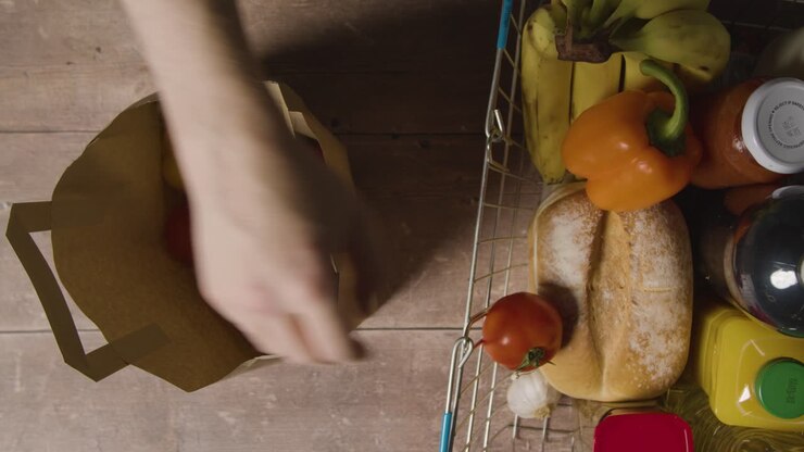 Overhead Shot Of Person Taking Basic Fresh Food Items From Paper Bag And Putting Them Into Supermarket Shopping Basket