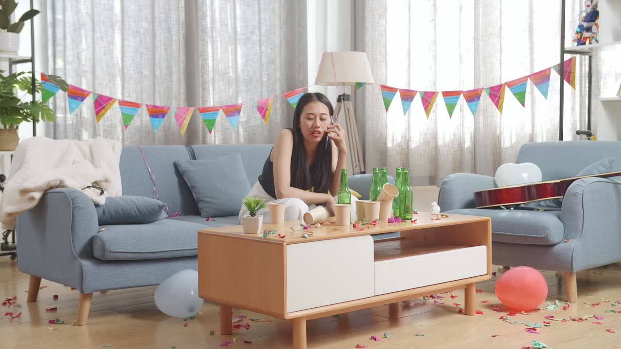 술에 취한 아시아 여성이 소파에 앉아서 집에서 파티를 한 후 스마트폰으로 이야기하고 있습니다.
