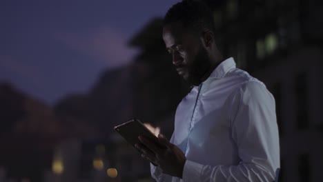 Video-De-Un-Reflexivo-Hombre-De-Negocios-Afroamericano-Usando-Una-Tableta-Por-La-Noche-En-La-Terraza-De-La-Oficina