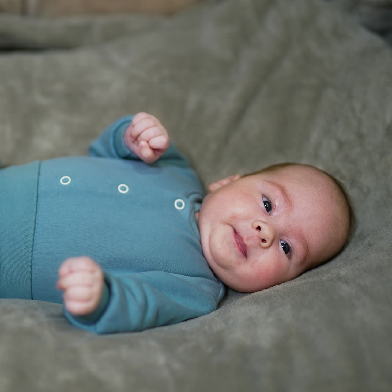 Healthy beautiful toddler lies on the bed and tosses his limbs. Adorable baby boy in warm blue costume on the grey background