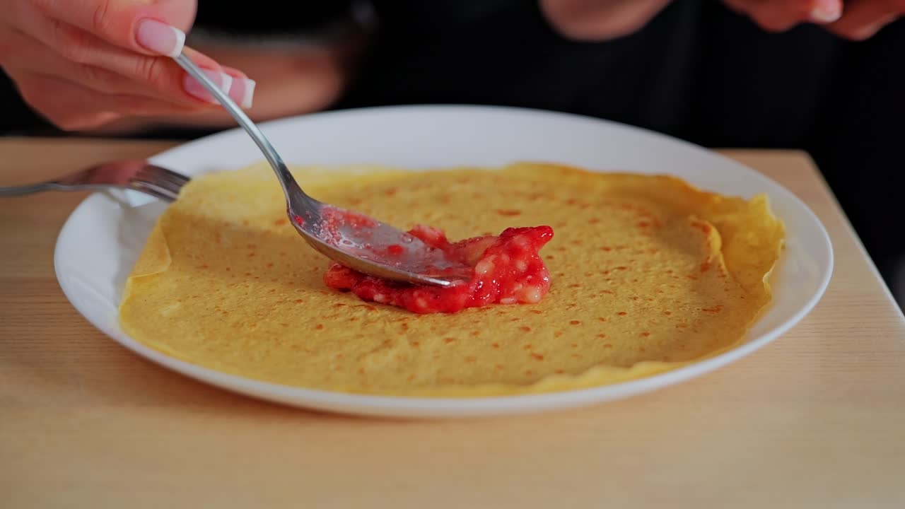 Latvian woman adds cold strawberry blend on crepe for breakfast on wooden table