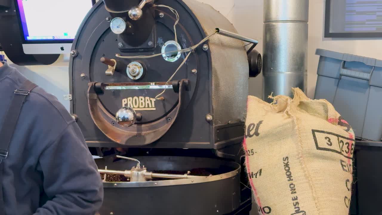 A coffee roasting machine operates in a cafe, surrounded by sacks of beans. Warm lighting enhances the industrial setting