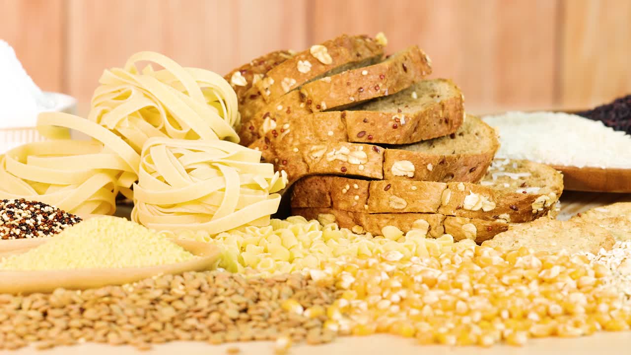 Whole wheat bread, pasta, and grains arranged on a wooden background with warm lighting and static camera