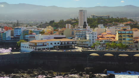 Tourists-walking-in-the-harbor-of-Puerto-de-la-Cruz,-Tenerife,-Canary-Islands,-its-stone-piers,-rocks-and-reefs-washed-by-the-waves-of-the-Atlantic-ocean-on-a-sunny-summer-day,-zooming-aerial-shot-4K