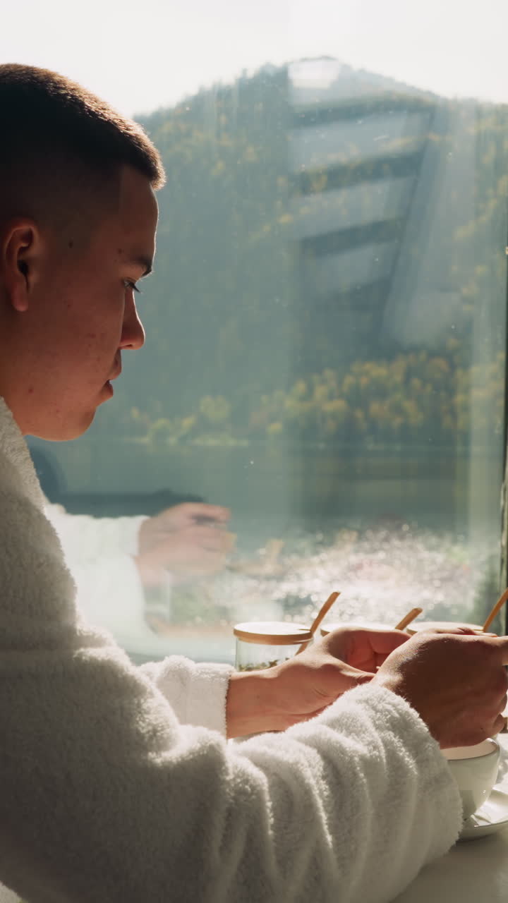 한 남자가 창문 에 앉아 차를 준비하고 있다. 집안 옷을 입은 집중된 남자가 주방에서 술을 마시기 위해 항아리에서 건조한 잎을 가져간다. 아침 의식