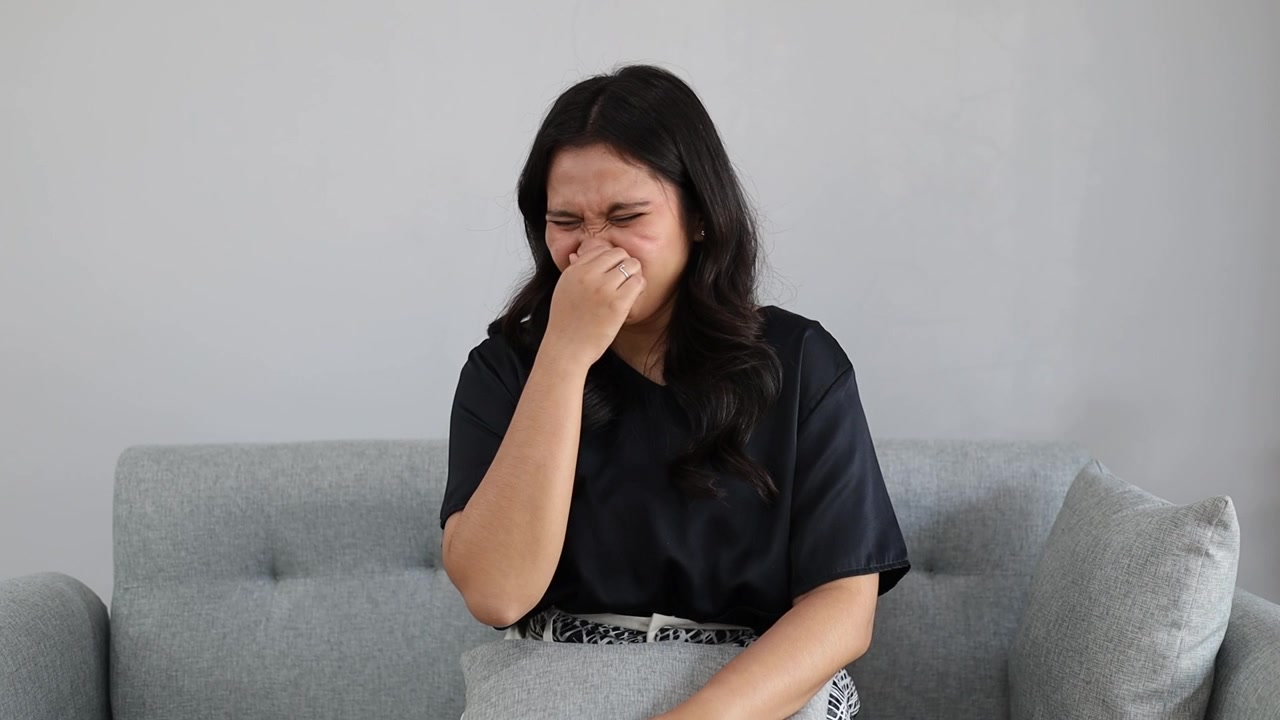 Giovane donna asiatica attraente seduta sul divano di casa che annusa qualcosa di puzzolente e disgustoso, odore intollerabile, trattenendo il respiro con le dita sul naso. Cattivo odore