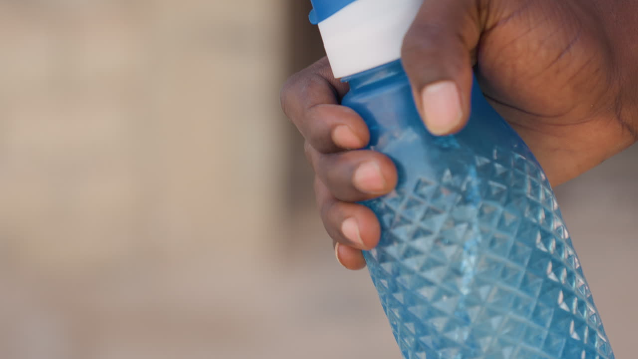 Zwarte hardloper knijpt in een blauwe sportfles; close-up van de getextureerde grip en de duim die de klapdop indrukt, handpalm detail met bezwete vingers, energie van het parcours en de baan, verfrissing halverwege de training, fietser en trainer.