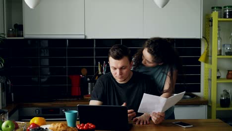 sad unhappy young man worried about bills payment using laptop in the kithcen at home. his girlfrend comes and support him