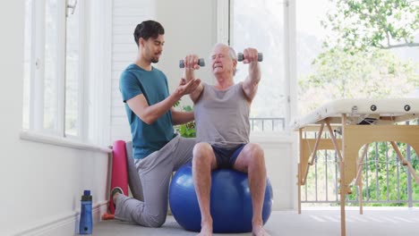 Männlicher-Trainer-Kniet-Neben-Einem-älteren-Mann,-Der-Im-Fitnessstudio-Mit-Hanteln-Auf-Einem-Fitnessball-Trainiert