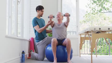instructor masculino arrodillado por un hombre mayor haciendo ejercicio con pesas en una pelota de fitness en el gimnasio