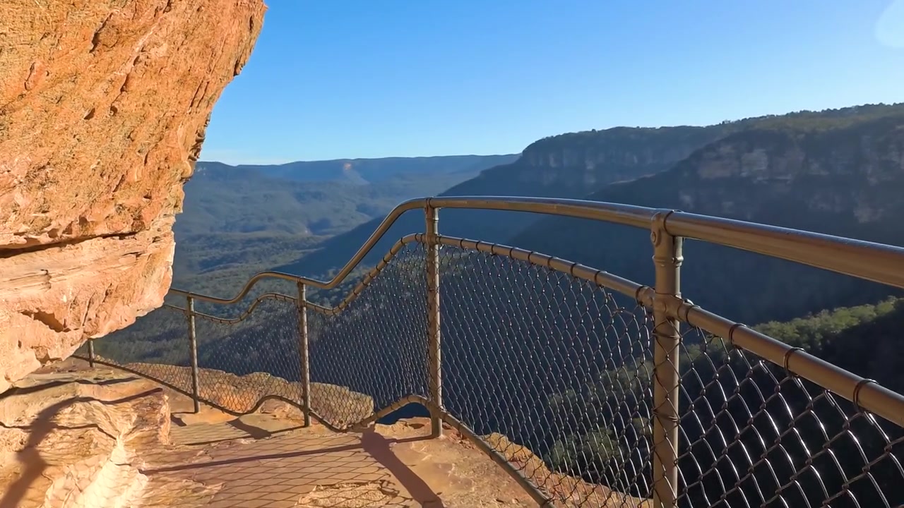 A scenic sandstone cliff path with handrails overlooks a vast forested canyon in Australia.