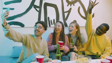 divertidos amigos jóvenes multiétnicos tomando un video selfie mientras se sientan en el sofá y tienen una fiesta de cerveza en casa
