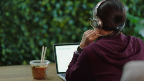 rear view young woman student using laptop computer browsing online working on project listening to music wearing headphones in modern office