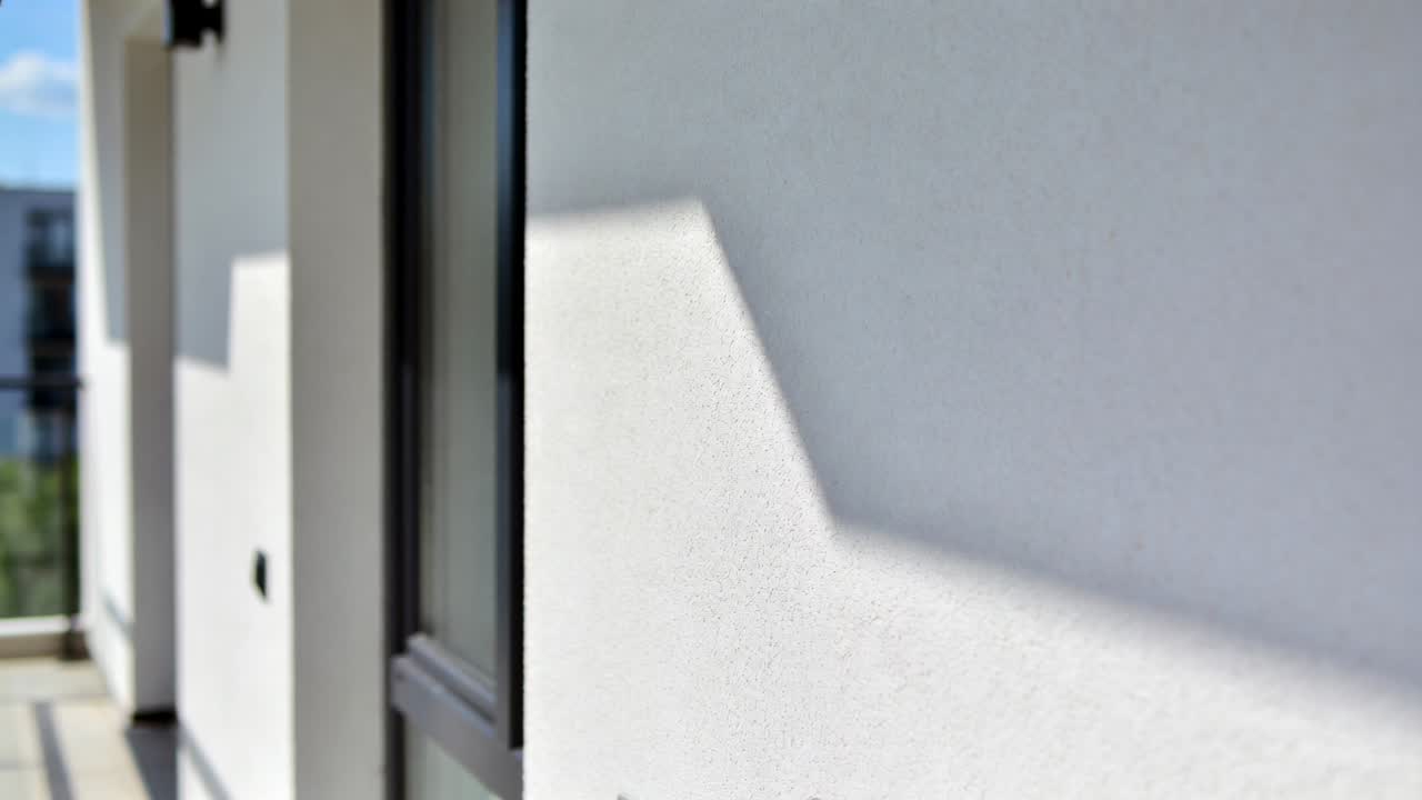 Luz solar cambiante, pared de estuco blanca mostrando una sombra escalonada en un balcón de apartamento con barandilla de cristal.