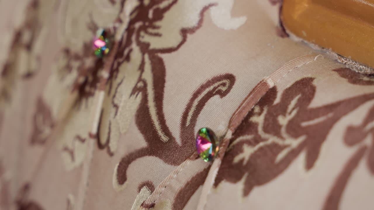 Primo piano di tappezzeria capitonné con bottone gioiello su tessuto floreale vintage, toni beige e marroni, texture cucita, piccoli strass che riflettono la luce, bordo sottile della cornice in legno che suggerisce la falegnameria.