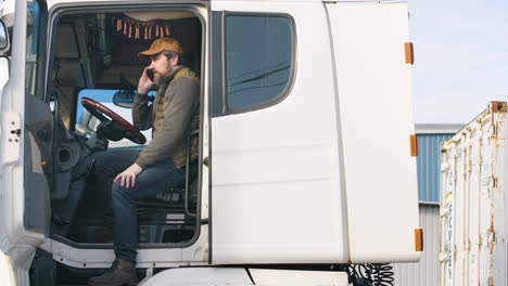 caucasian worker wearing vest and cap organizing a truck fleet in a logistics park while talking on the phone in a truck