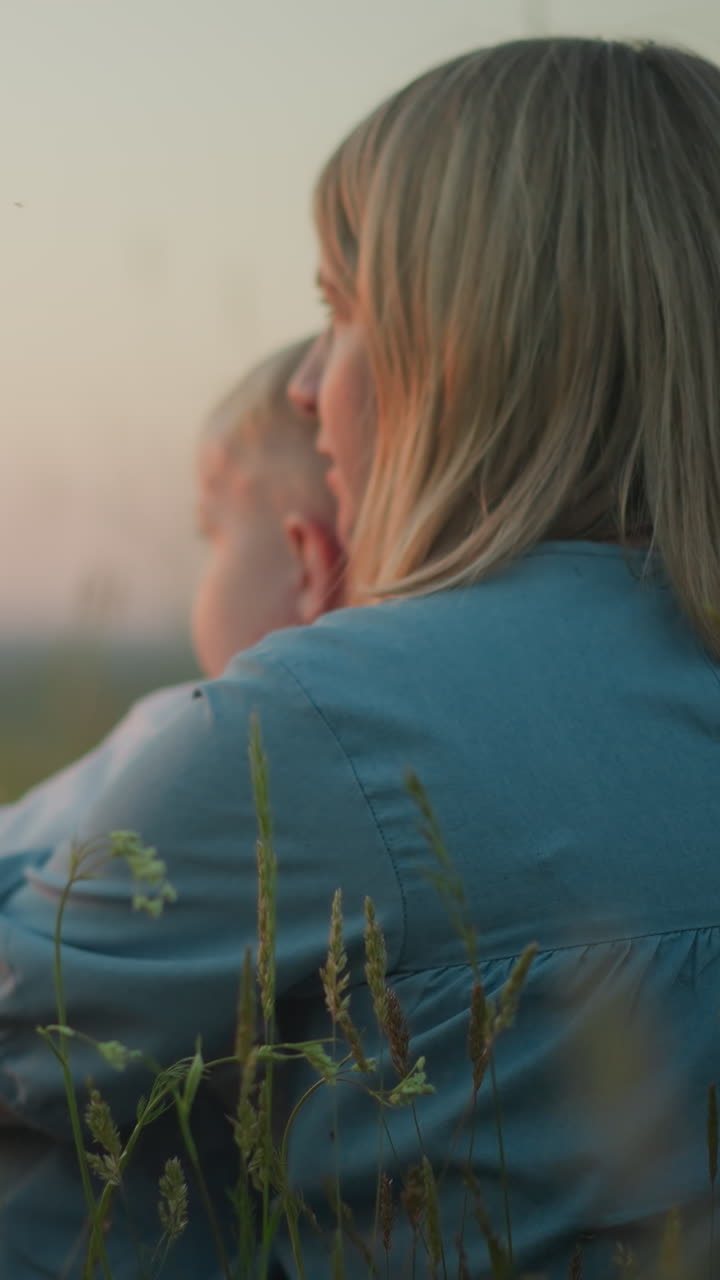 해가 지는 동안 들판에 앉아있는 파란 옷을 입은 어머니의 클로즈업 백, 어린 아이를 무에 들고 부드럽게 움직이고, 큰 아이는 모두 호수를 바라보면서 에 앉습니다.