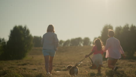 family enjoying outdoor activity with dog during sunny day, woman in light blue shorts holding flowers while child runs alongside with dog on leash