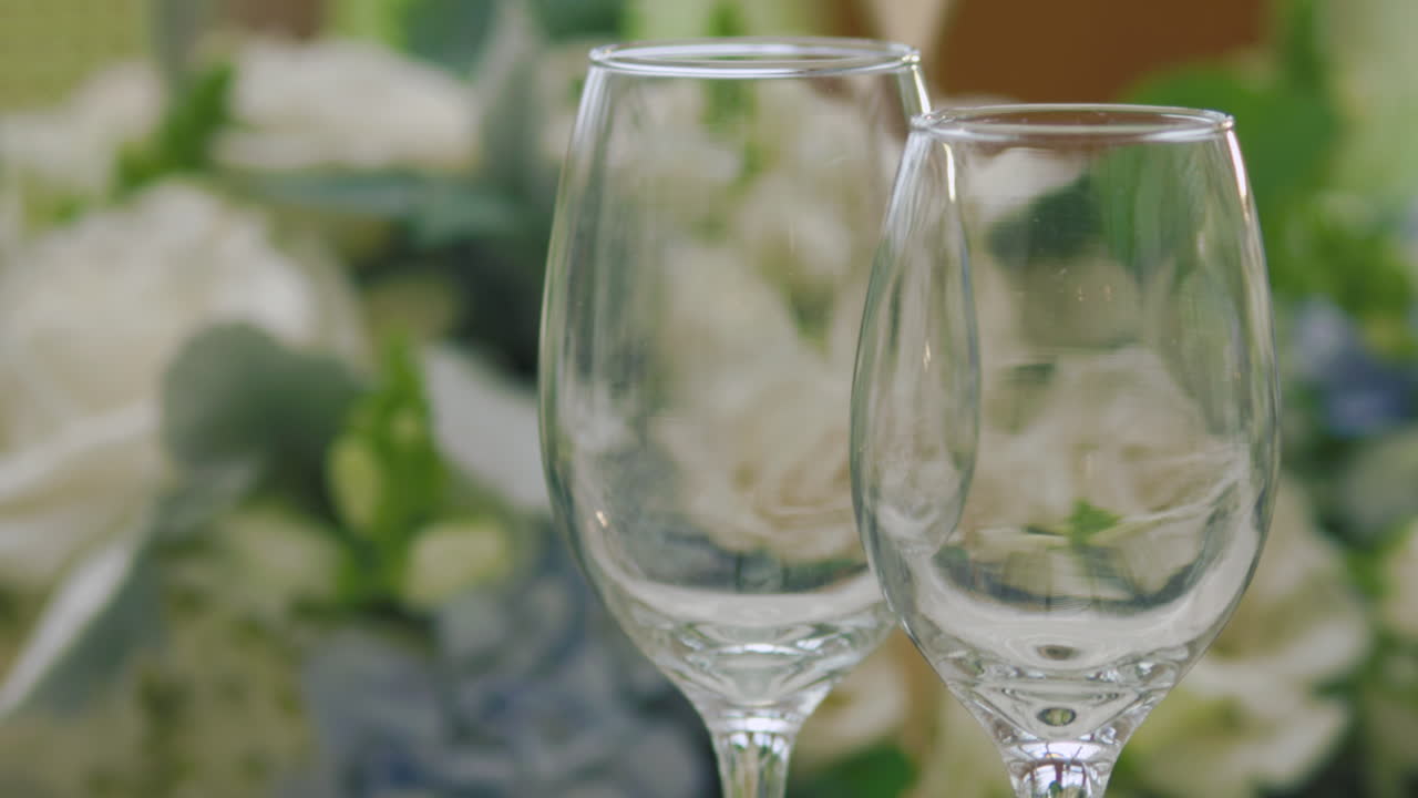 An elegant visual detail where the initial focus shows glass cups on the table and, through gentle panning, it blurs to focus on a delicate floral arrangement.
