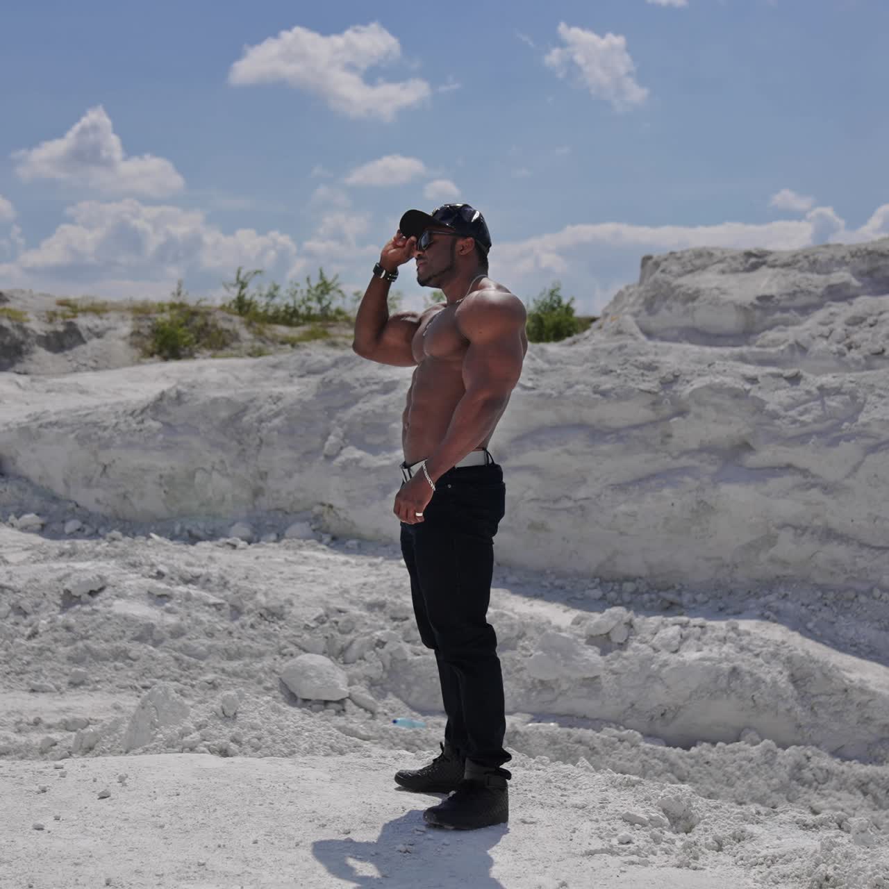 Handsome guy without shirt standing on white rocky background. Dark skinned guy in black cap and glasses with muscular body outdoors. Profile view