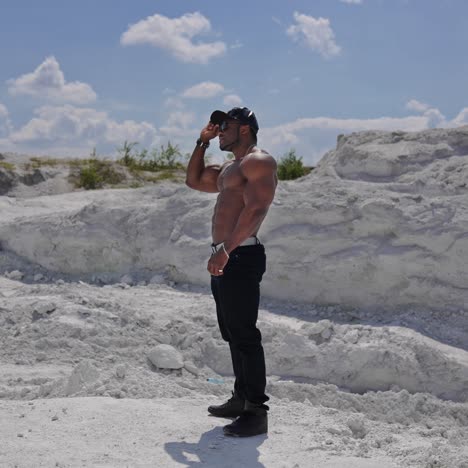 handsome guy without shirt standing on white rocky background. dark skinned guy in black cap and glasses with muscular body outdoors. profile view