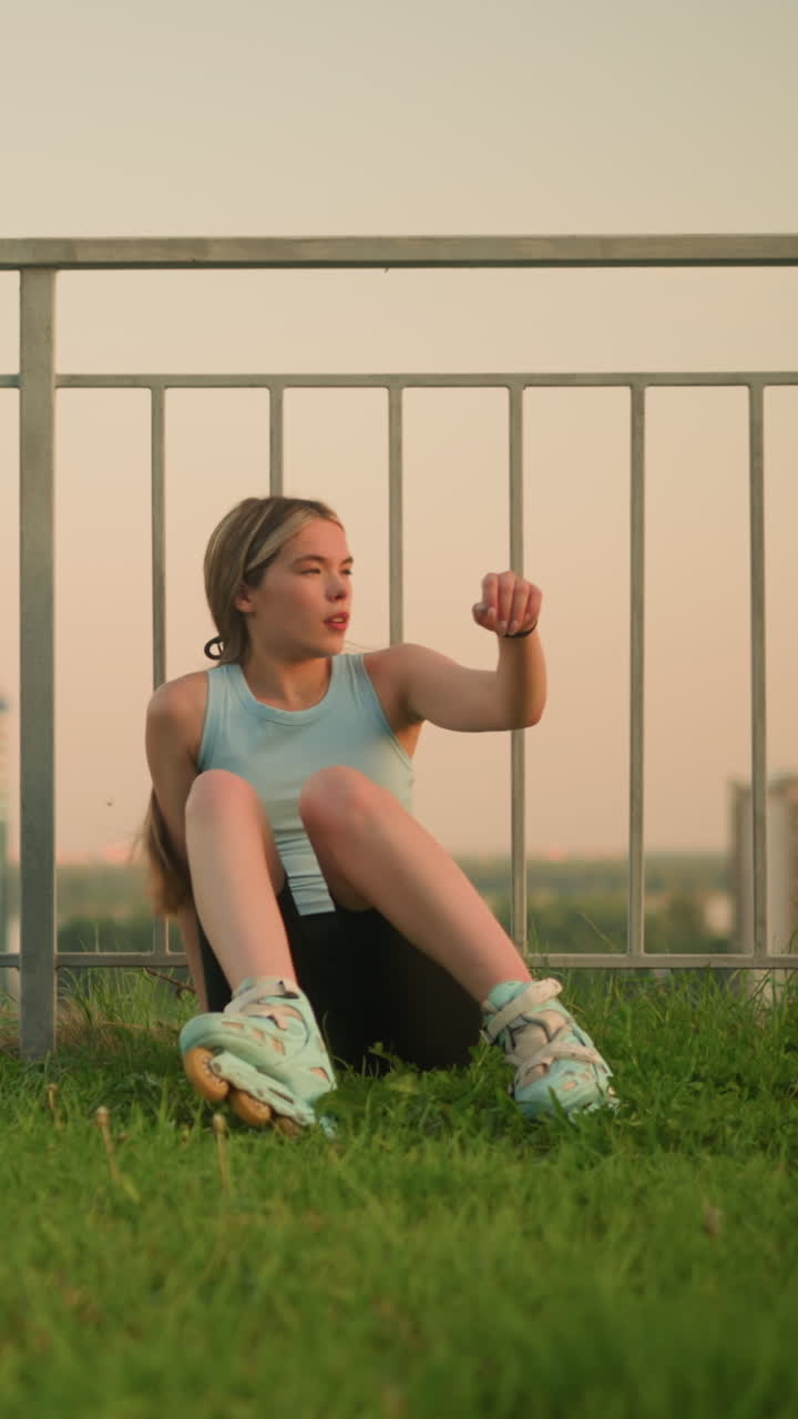 joven en patines sentada en un terreno cubierto de hierba, descansando con la mano en la pierna, disfrutando de la puesta de sol nocturna con el paisaje urbano borroso en el fondo