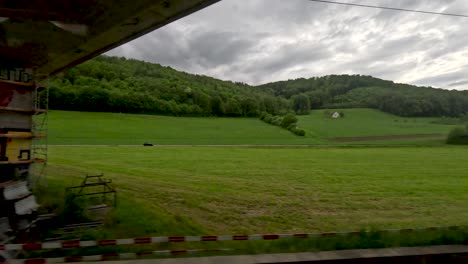 Blick-Aus-Dem-Zugfenster,-Während-Der-Zug-An-Ländlichen-Grünen-Feldern-Mit-Bewaldeten-Sanften-Hügeln-Unter-Einer-Neuen,-Im-Bau-Befindlichen-Brücke-Vorbeifuhr