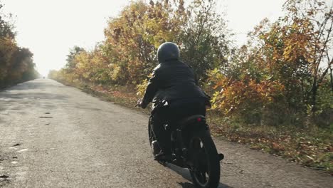 Seiten--Und-Rückansicht-Eines-Mannes-Mit-Schwarzem-Helm-Und-Lederjacke,-Der-Im-Herbst-Auf-Einer-Asphaltstraße-Motorrad-Fährt.-Bäume-Mit-Braun