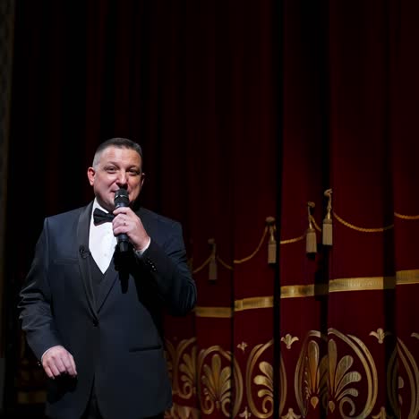 speaker on stage. male host in elegant costume speaking into a microphone looking on camera on the background of red curtains in the theater. slow motion