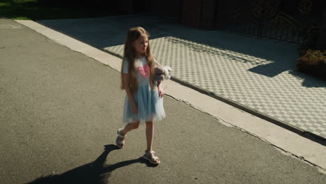 young girl with long hair and blue dress walks casually under sunlight holding stuffed animal, casting clear shadow on paved sidewalk next to checkered pavement