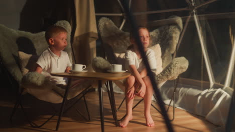 children sit at table in glamping tent. pensive little boy with girl sit tired in cosy chairs by transparent wall. tranquil kids spend time on terrace at night