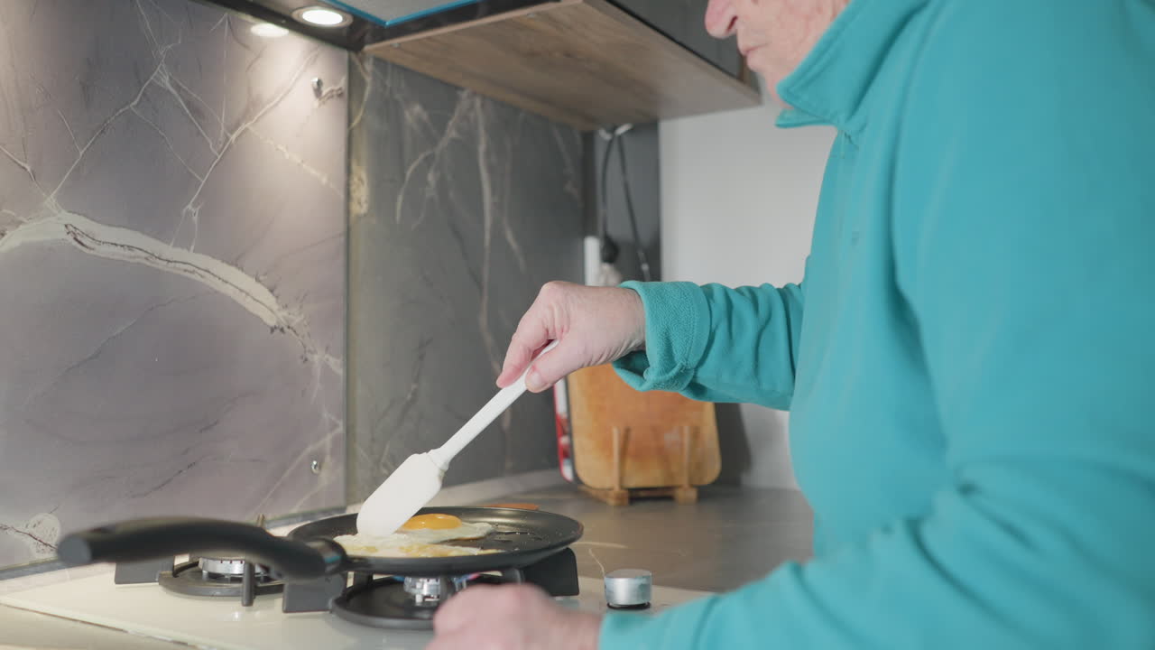 Older woman flipping eggs in frying pan with spatula, cooking on stove in modern kitchen, focused on food preparation process with kitchen tools, stovetop, and meal visible