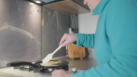 older woman flipping eggs in frying pan with spatula, cooking on stove in modern kitchen, focused on food preparation process with kitchen tools, stovetop, and meal visible