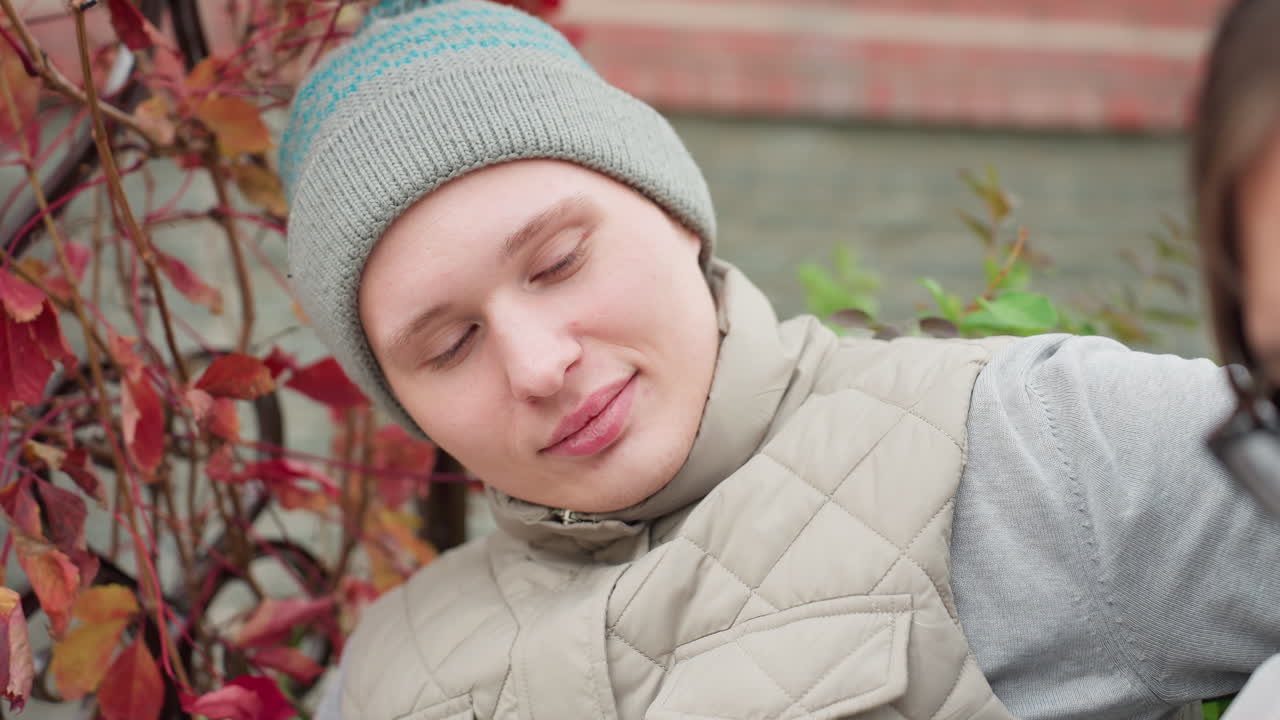 Close up of young man seated outdoors in autumn wearing knit cap and quilted jacket, soft expression on face as he hug his wife, red and orange leaves in background