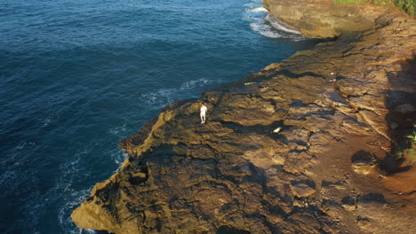 epic adventure seeker drone shot of urban nomad traveler, hipster young millennial man stand on edge of cliff in indonesia, confident and brave, search for new exciting horizons and ideas